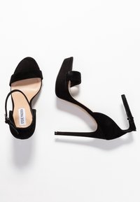 Sandales à talons hauts en daim noir avec brides à la cheville, orteils ouverts et talons fins sur fond blanc, une chaussure debout et une couchée sur le côté.