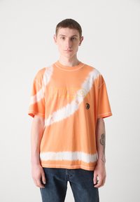 Oranžinis tye-dye medvilninis marškinėliai su dideliu geltonu "hugo" logotipu ir maža juoda grafika, pasižymintys laisvu kirpimu ir trumpomis rankovėmis.