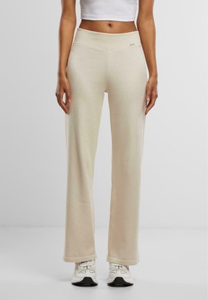 Beige, gerippte Lounge-Hosen mit hohem Bund, die eine glatte Textur und eine lockere Passform bieten, kombiniert mit weißen Sportschuhen.