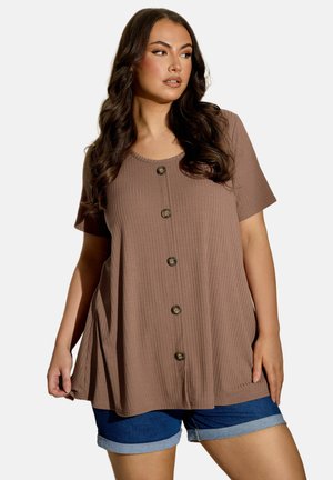 Kvinde med langt mørkt hår iført en løs, kortærmet brun knapbluse og op-rullede jeansshorts, der kigger til siden.