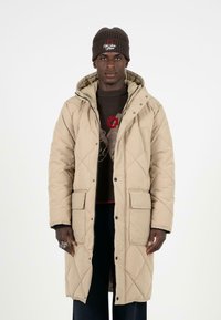 Béžový prošívaný dlouhý kabát s patentkami, dvěma předními kapsami a kapucí. Nosený přes hnědý svetr se složitým vzorem.