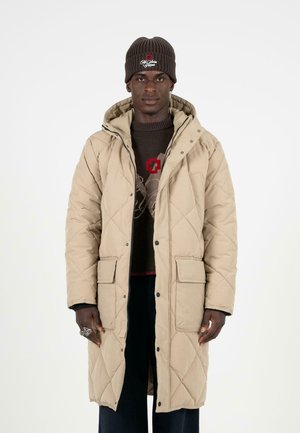 Beige gesteppter langer Mantel mit Druckknöpfen, zwei vorderen Taschen und Kapuze. Getragen über einem braunen Pullover mit einem komplizierten Muster.