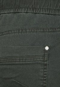 Un tejido de algodón verde oscuro presenta un bolsillo trasero visible con un broche plateado. La textura es suave con costuras cosidas para mayor durabilidad.