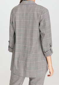 Femme portant un blazer à carreaux gris avec les manches retroussées et un pantalon à carreaux assorti, vue de dos sur un fond uni.