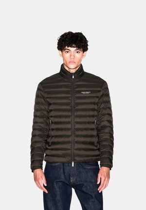 Joven con pelo rizado que lleva una chaqueta acolchada negra de Armani Exchange cerrada con cremallera, combinada con jeans azul oscuro, sobre un fondo liso.