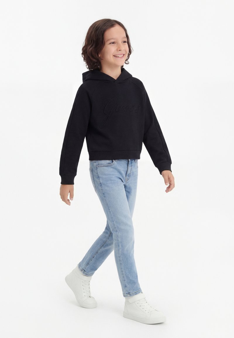 Copil cu păr creț care merge pe jos, purtând un hanorac negru, blugi albastru deschis și adidași albi, zâmbind și privindu-se în lateral.