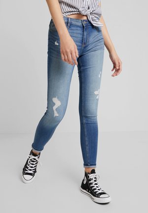 Femme portant un jean skinny bleu déchiré et des baskets montantes noires marchant sur un sol gris avec un fond gris uni.
