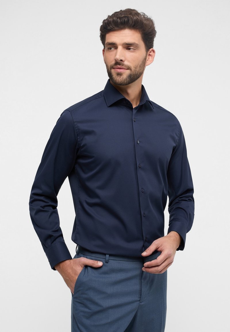 Herren Langarmhemd aus dunkelblauem Stoff, strukturierte Gestaltung mit klassischem Kragen, Frontknöpfen und glatter Textur. Kombiniert mit hellblauen Hosen.
