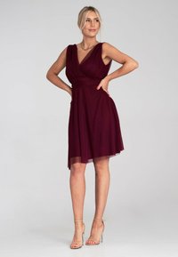 Burgunderfarbene ärmellose Kleid mit V-Ausschnitt, gerafftem Bund und fließendem Rock. Kombiniert mit transparenten High Heels. Glatte Stoffoberfläche.