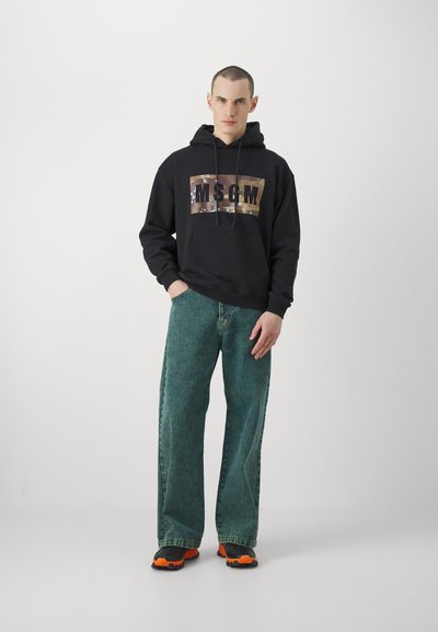 Schwarzer Hoodie mit einem Camouflage-Graphic und "MSGM"-Text, kombiniert mit weit geschnittenen, ausgewaschenen grünen Jeans und schwarz-orangefarbene Sneakers. Minimalistischer Hintergrund.