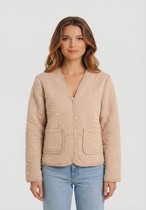 Mujer con chaqueta acolchada beige con motivos de hojas y dos bolsillos frontales, combinada con jeans azul claro, posando frente a un fondo liso.