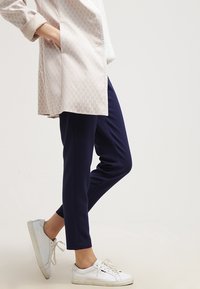 Lichtroze, gebloemde lange jas met geometrisch ontwerp, gecombineerd met strakke marineblauwe broeken en witte sneakers. Eenvoudige, moderne en casual uitstraling.