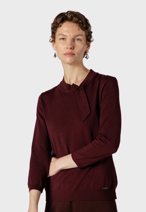 Femme aux cheveux courts et bouclés portant un haut bordeaux à manches longues avec un détail de nœud au cou et des boucles d'oreilles géométriques pendantes sur un fond uni.