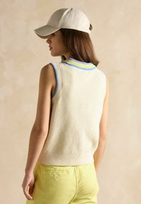 Beige spalvų megztinis be rankovių su tekstūruotu audiniu, mėlynai-geltonos juostelės apykakle. Suderintas su šviesiai geltonomis kelnėmis ir smėlio spalvos kepure.