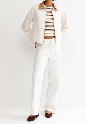 Person trägt weiße Jacke mit beige-farbenem Kragen, gestreiftes Crop-Top, weiße Hose und braune flache Schuhe, steht mit den Händen in den Taschen.
