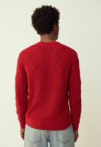 Piros kötött pulóver texturált hullámmintával, bordázott mandzsettával és szegéllyel, világoskék farmerrel viselve, hátulnézetből.