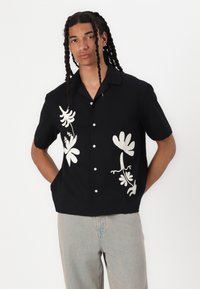 Zwart shirt met korte mouwen en witte botanische prints; beschikt over een kraag en knopen aan de voorkant, gemaakt van lichte stof en biedt een ontspannen pasvorm.