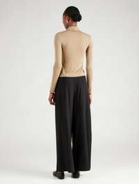 Beige Bluse mit langen Ärmeln und hohem Kragen, kombiniert mit weiten schwarzen Hosen, von hinten betrachtet. Flache schwarze Schuhe.