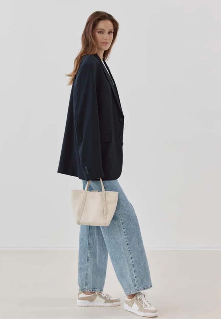 Overmaatse zwarte blazer, lichtblauwe wijde jeans, beige handtas, witte sneakers met subtiele bruine accenten, gladde textuur en strakke lijnen.