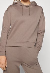 Sudadera con capucha marrón hecha de tela suave, que cuenta con un bolsillo frontal tipo canguro, capucha con cordón y puños y dobladillo acanalados.