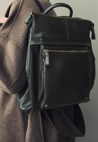 Sac à dos en cuir noir avec une finition texturée, poignée supérieure, poche avant zippée et accents de quincaillerie dorée. Porté sur un vêtement en tissu marron.