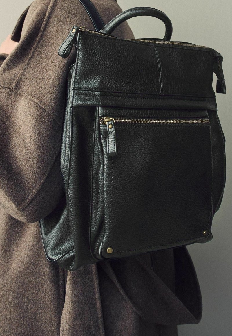 Sac à dos en cuir noir avec une finition texturée, poignée supérieure, poche avant zippée et accents de quincaillerie dorée. Porté sur un vêtement en tissu marron.