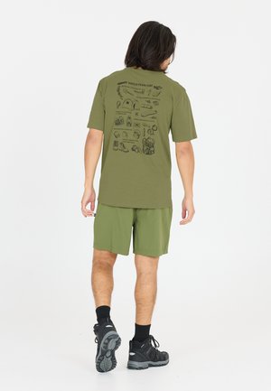 Man draagt een olijfgroen T-shirt met illustraties van kampeeruitrusting op de achterkant, bijpassende korte broek, zwarte sokken en zwarte wandelschoenen terwijl hij wegloopt.