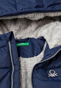 Chaqueta tipo puffer en color navy con un suave forro de felpa gris, que cuenta con un cierre de cremallera y un parche con el logo. La etiqueta interior dice "United Colors of Benetton."