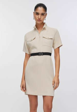 Donna che indossa un abito beige a maniche corte con tasche frontali, collo con mezza cerniera e cintura nera in vita.
