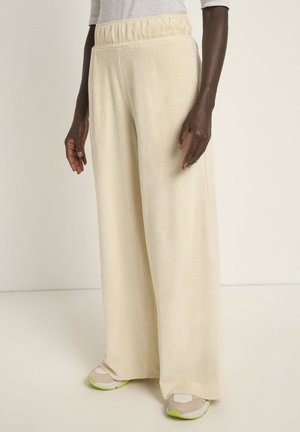 Wide-leg beige pants in soft fabric with an elastic waistband. Notable texture and loose fit enhance comfort. Paired with neutral sneakers.