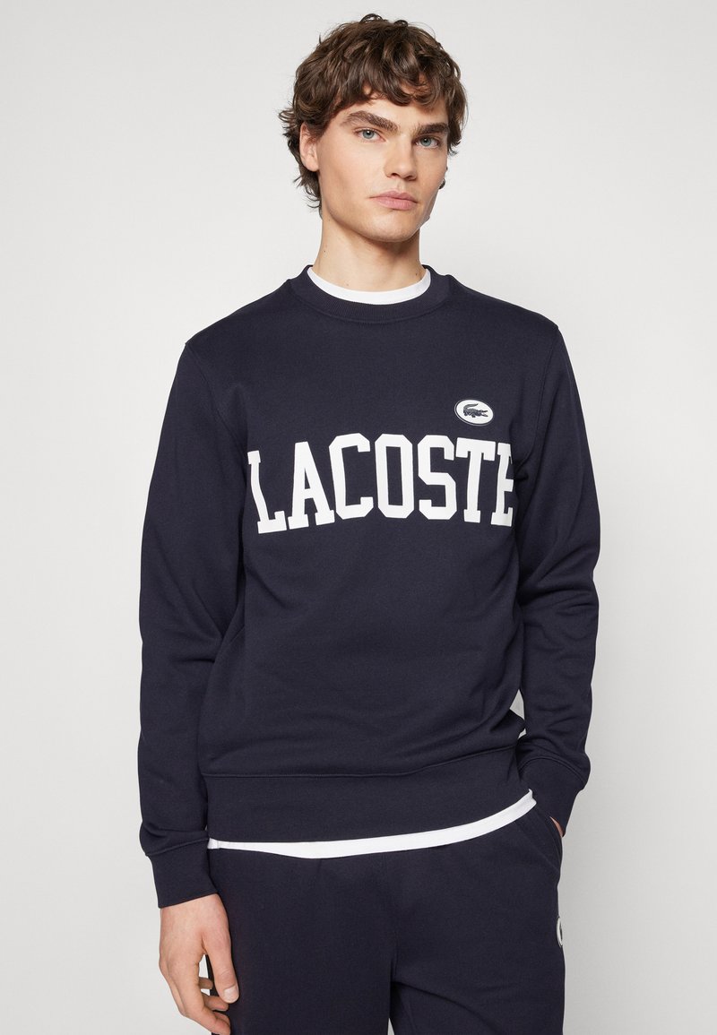 Sudadera azul marino de tela suave, con letras blancas "LACOSTE" y un parche del logo en el pecho. Cuello redondo y puños acanalados.