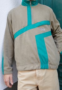 Fleece pullover i beige med teal detaljer, der har en halv lynlås i halsen, kontrasterende paneldesign og elastisk kant og manchetter.