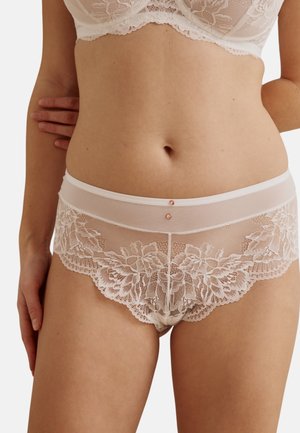 Femme portant un soutien-gorge en dentelle blanche et un slip assorti en dentelle florale, debout avec une main reposant sur le côté.