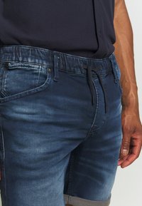 Tummansiniset farkkushortsit, joissa on joustava vyötärö ja nyöri, etutaskut ja haalistunut pinta. Lahkeiden hupit ovat käännetty vaaleammalla sävyllä.