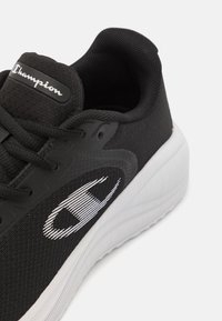 Svart träningsskor med övre del i mesh, vit gummisula och framträdande vitt Champion-logotyp. Har en texturerad design på materialet.