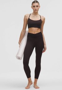 Top scurt negru cu bretele subțiri și colanți negri cu talie înaltă, evidențiind un design texturat, ținut cu un covor de yoga deschis la culoare, rulat.