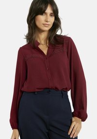 Vaarikapunane pikkade varrukatega pluus, ees nööpidega, pitsdetailidega rinna piirkonnas ja lõõgastunud lõikega, kombineeritud tumesiniste pükstega.