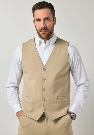 Homme barbu portant un gilet beige boutonné sur une chemise blanche, pantalon beige, debout les mains dans les poches devant un fond uni.