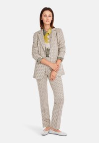 Patterned suit in cream and grey with geometric design. Collared floral shirt underneath. White flat shoes with decorative accents.