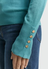 Der türkisfarbene Pullover verfügt über gerippte Bündchen mit drei goldfarbenen Knöpfen, eine glatte Textur und eine körperbetonte Passform, kombiniert mit blauem Denim.