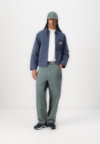 Jachetă albastră din denim, tricou alb, blugi verzi cu croială largă și adidași negri. Căciula este verde cu text alb. O ținută simplă și casual.