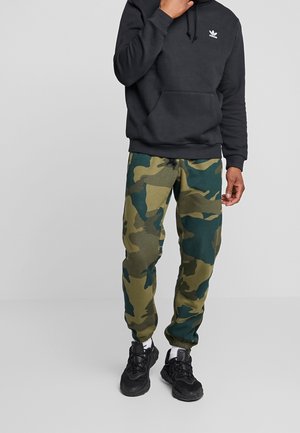 Jogging en camouflage vert et bleu avec une taille élastique et un cordon de serrage. Porté avec un hoodie noir et des baskets noires.