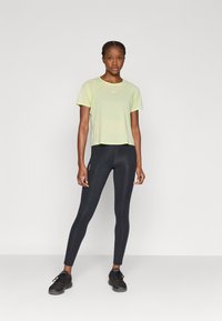 Αθλητική κοντομάνικη μπλούζα σε ανοιχτό πράσινο χρώμα με αντανακλαστικές λεπτομέρειες, συνδυασμένη με μαύρο κολάν και μαύρα αθλητικά παπούτσια.