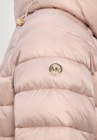 Manga de chaqueta acolchada rosa claro con insignia circular dorada del logo MK de Michael Kors adjunta cerca del hombro.