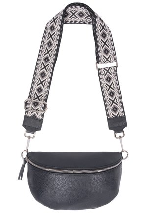 Femme portant un crop top beige et un short vert avec un sac bandoulière en cuir noir présentant une sangle réglable à motif.