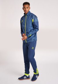 Herrarnas sportklädes-set inkluderar en marinblå jacka med neonlysande detaljer, andningsbart material och matchande marinblå byxor med sidopaneler och logotyp.