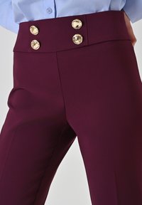 Pantalon bordeaux taille haute avec une texture lisse, des boutons double boutonnage dorés et une coupe ajustée.