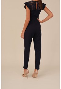 Marinblå jumpsuit med spetsärmar och detalj med nyckelhål bak. Tight passform med avsmalnande ben, i kombination med nude högklackade skor.