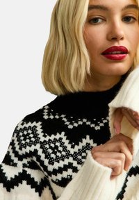 Knit sweater featuring a black and white geometric pattern, turtleneck design, and ribbed cuffs. Adorned with decorative beads on the chest.