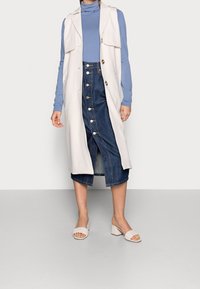 Gilet lungo beige con chiusura a bottone sopra un dolcevita blu, abbinato a una gonna di jeans con spacco frontale e bottoni, e sandali bianchi con tacco bloccato.
